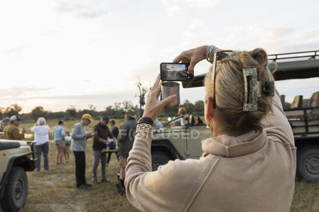 Rear view of woman taking smart phone picture of a group of people on an early morning game drive — Stock Photo