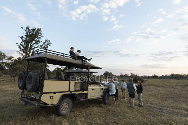 Grupo de personas de pie alrededor de vehículos de safari en una unidad de juego temprano en la mañana - foto de stock