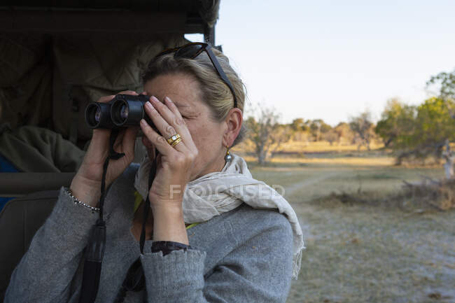 Adult woman looking through binoculars standing by a jeep. — Stock Photo