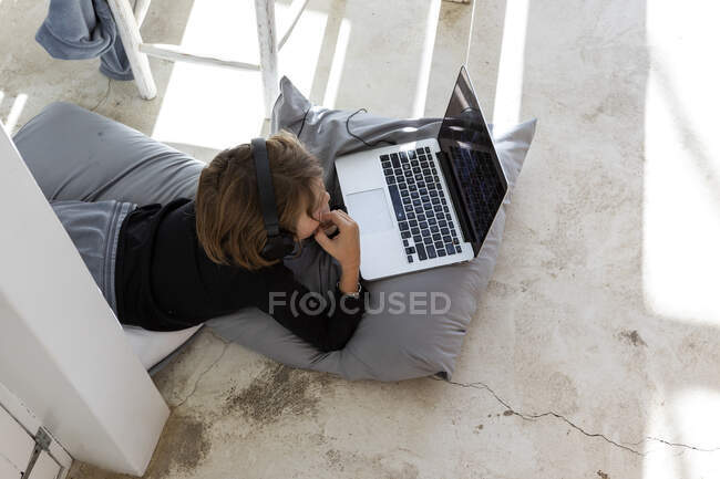 Achtjähriger Junge liegt auf Kissen, Kinn auf Händen, schaut auf Laptop-Bildschirm, macht Hausaufgaben. — Stockfoto