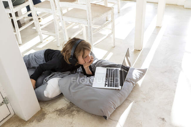 Niño de ocho años acostado en el suelo sobre cojines usando un portátil - foto de stock