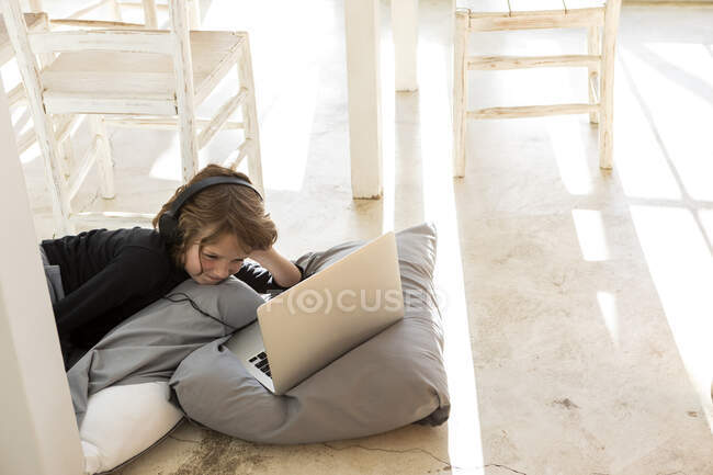 Otto anni ragazzo sdraiato sul pavimento su cuscini utilizzando un computer portatile — Foto stock