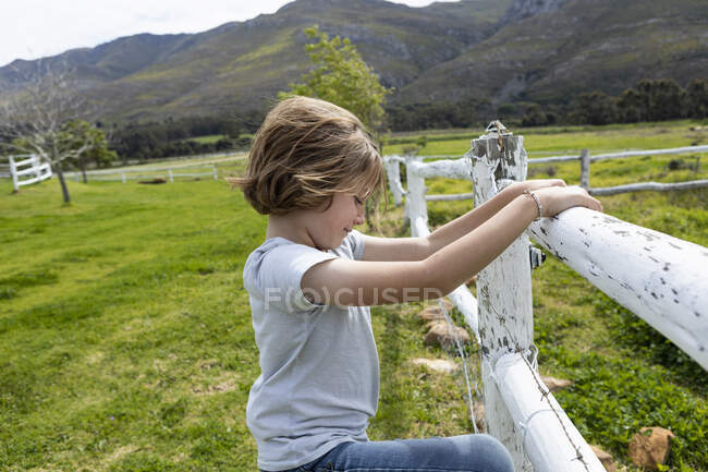 Menino de oito anos apoiado em uma cerca, olhando para cavalos em um campo — Fotografia de Stock
