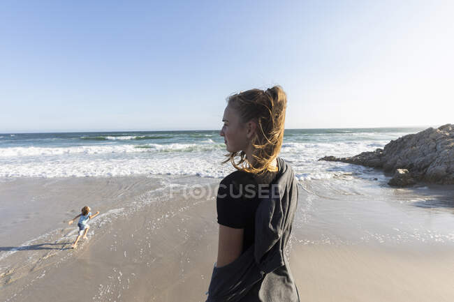 Adolescente de pie mirando hacia una playa, un chico corriendo en la arena debajo - foto de stock