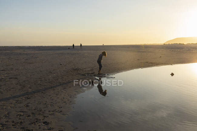 Garçon sur une plage, regardant son reflet dans une piscine d'eau, marée basse, coucher de soleil. — Photo de stock