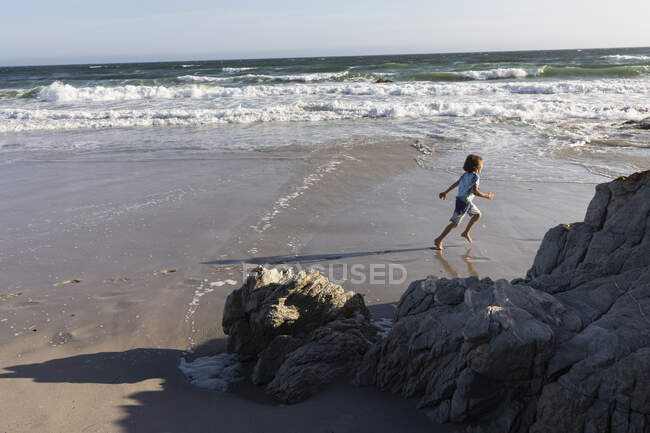 Un niño corriendo en la arena en el borde del agua en una playa de arena. - foto de stock