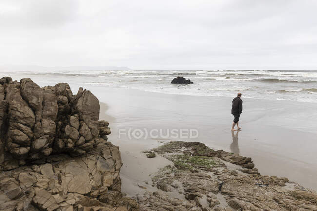 Un uomo che cammina attraverso la sabbia fino al bordo dell'acqua su una spiaggia, giorno coperto e onde da surf che si infrangono sulla riva. — Foto stock