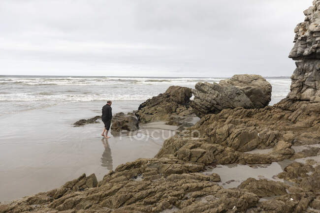 Чоловік, що йде через пісок до краю води на пляжі, похмурий день і хвилі серфінгу, що ламаються на березі . — стокове фото