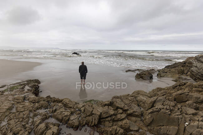 Un homme marchant à travers le sable jusqu'au bord de l'eau sur une plage, jour couvert et vagues de surf se brisant sur le rivage. — Photo de stock