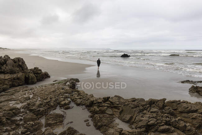 Un hombre caminando a través de la arena hasta el borde del agua en una playa, día nublado y olas de surf rompiendo en la orilla. - foto de stock