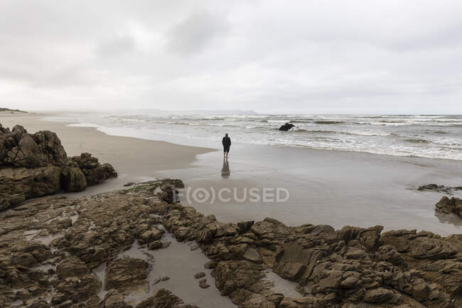 Um homem caminhando através da areia até a borda da água em uma praia, dia nublado e ondas de surf quebrando na costa. — Fotografia de Stock