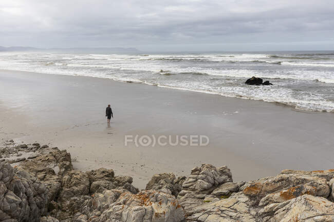 Ein Mann, der an einem Strand über Sand bis an den Rand des Wassers geht, trübt den Tag, und Brandungswellen brechen an Land. — Stockfoto