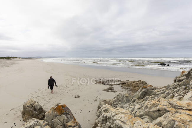 Un hombre maduro caminando por una playa llevando sus zapatos en la mano - foto de stock