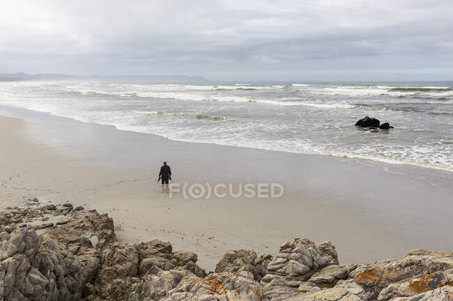 Человек, идущий по песку к краю воды на пляже, пасмурный день и волны для серфинга, разбивающиеся о берег. — стоковое фото