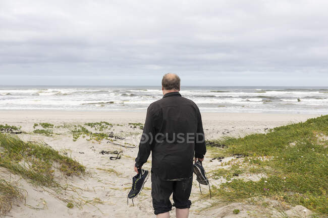 A mature man walking across a beach carrying his shoes in his hand — Stock Photo
