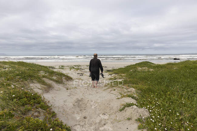 A mature man walking across a beach carrying his shoes in his hand — Stock Photo