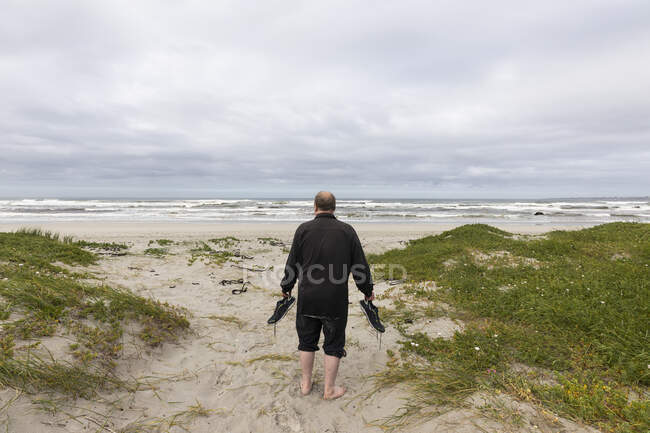 Зрілий чоловік, що йде через пляж, носить взуття в руці — стокове фото