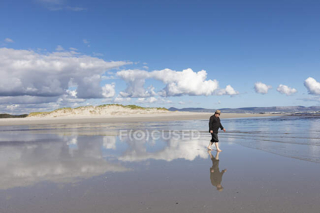 Un uomo adulto con un cappello che cammina lungo una spiaggia sabbiosa. — Foto stock
