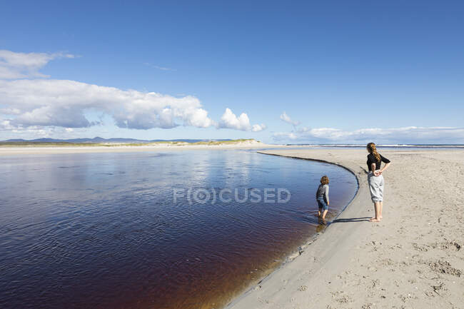 Teenage girl and a young boy at the edge of a wide water channel, on a sandy beach. — Stock Photo