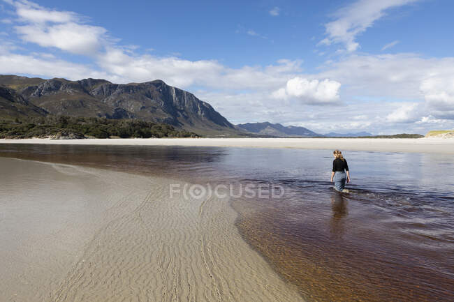 Ein Teenager-Mädchen watet durch einen Wasserkanal an einem breiten Sandstrand. — Stockfoto