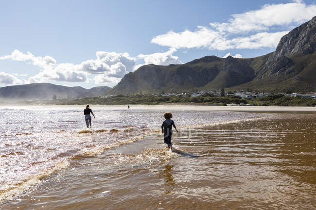 Adolescente y un niño corriendo a través de aguas poco profundas en una amplia playa de arena - foto de stock