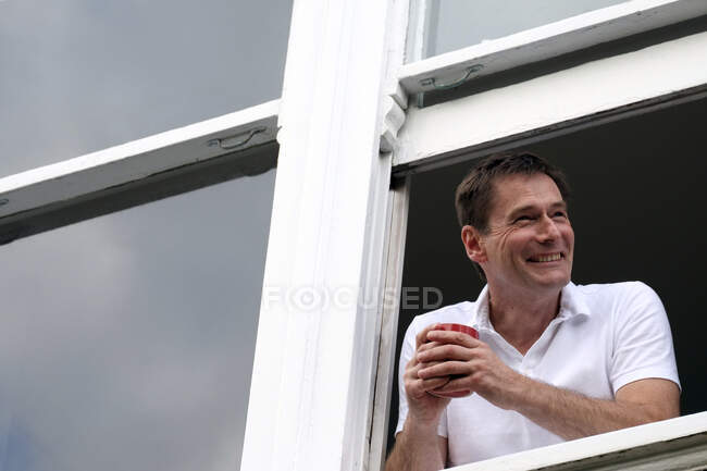 Smiling man leaning out of window holding hot drink. — Stockfoto