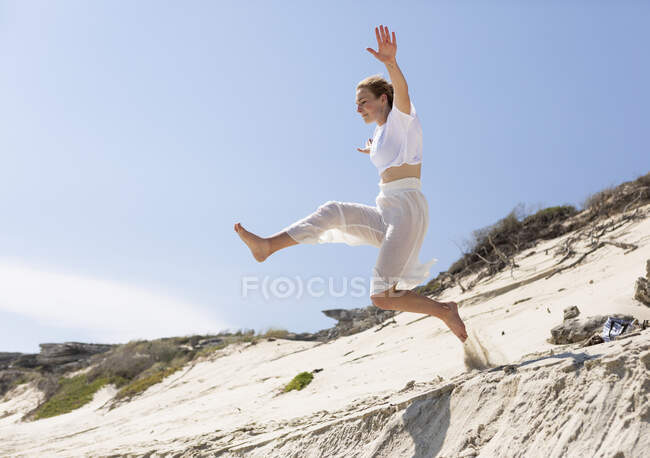 A teenage girl leaping from a sand dune into the soft sand below. — Stockfoto