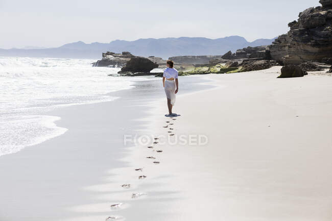 Adolescente caminando sola en una playa de arena en Sudáfrica por el borde del agua, vista trasera - foto de stock