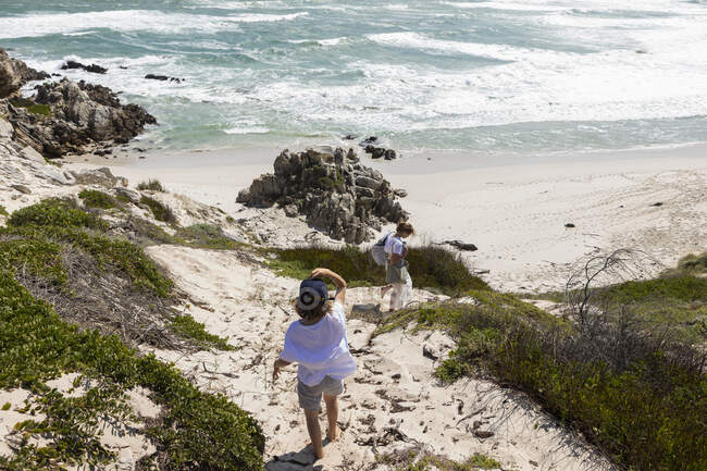 Adolescente y hermano menor con vistas a una playa y una costa rocosa con olas estrellándose en la orilla. - foto de stock