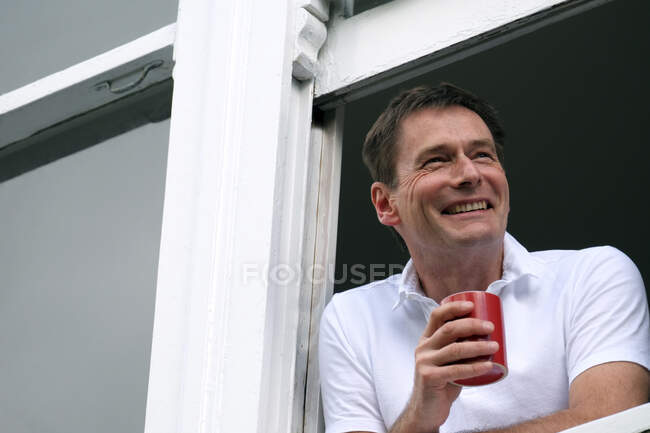 Smiling man leaning out of window holding hot drink. — Stockfoto
