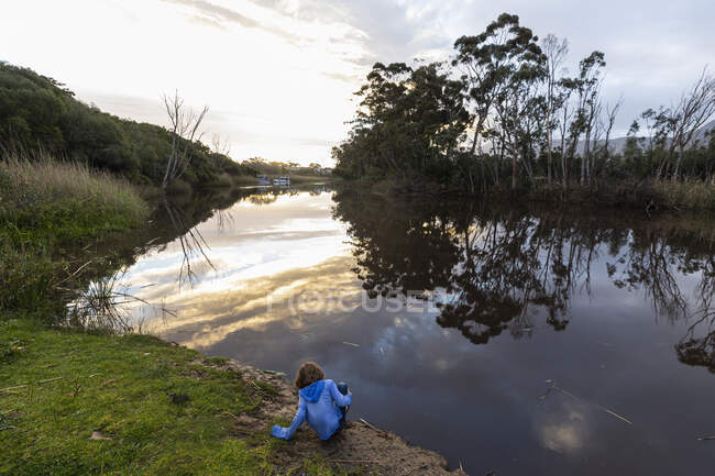 Um menino de pé junto a um rio ao entardecer, reflexos do céu na água calma plana — Fotografia de Stock
