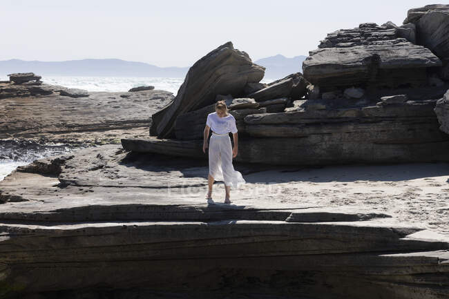 Ragazza adolescente arrampicata su rocce piatte lisce stratificate sopra una spiaggia sabbiosa con onde che si rompono sulla riva. — Foto stock