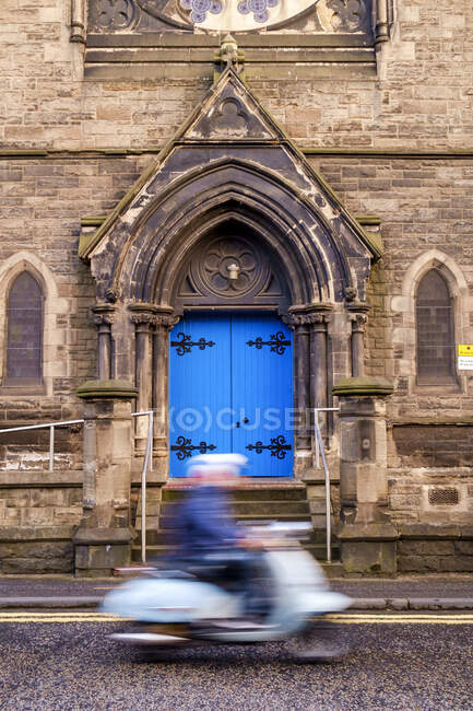 Person auf einem Motorroller fährt an einem gotischen Bogen mit hellblauer Tür vorbei. — Stockfoto