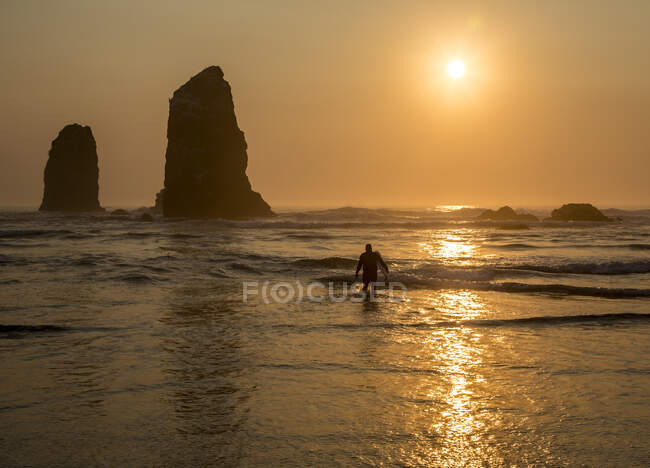 Серфер, выходящий из моря с заходящим солнцем за скалой в волнах на пляже. — стоковое фото