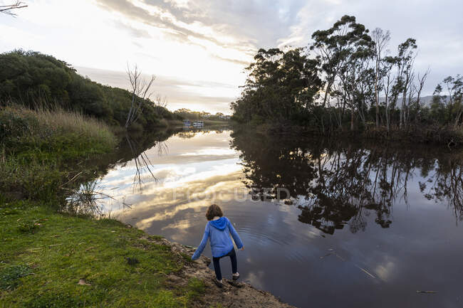 A young boy standing by a river at dusk, sky reflections in the flat calm water — Stockfoto
