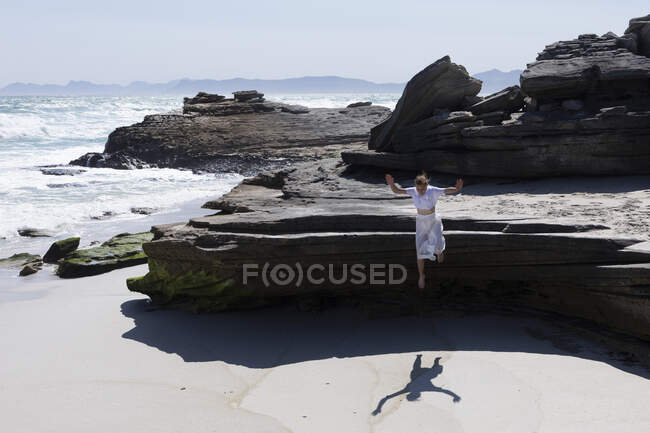 Teenage girl jumping onto sand from the top of smooth flat layered rocks above a sandy beach — Stockfoto