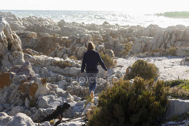 Adolescente grimpant sur les rochers déchiquetés sur une plage, de grandes vagues se brisant sur le rivage — Photo de stock