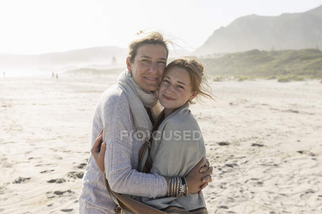 Mujer adulta y su hija adolescente abrazándose, de pie en una playa azotada por el viento - foto de stock