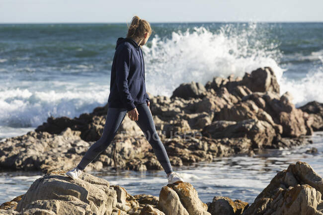 Teenage girl balancing on jagged rocks on a beach, surf breaking behind her. — Stock Photo