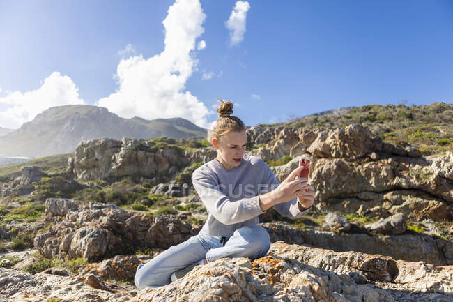 Девочка-подросток фотографирует со своего смартфона, сидит на скалах на пляже — стоковое фото