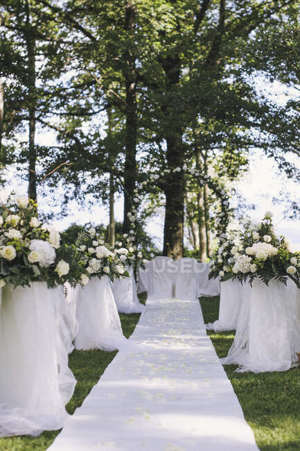 Un jardín con mesas a la sombra de árboles altos, un arco floral, escenario para una boda - foto de stock