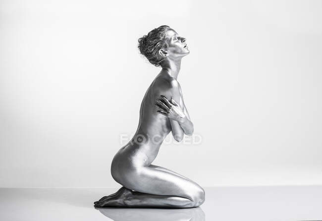 Mujer caucásica desnuda con pintura corporal plateada arrodillada, vista lateral, brazos cruzados a través de su cuerpo - foto de stock