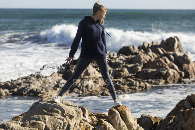 Adolescente equilibrio en rocas dentadas en una playa, surf rompiendo detrás de ella. - foto de stock