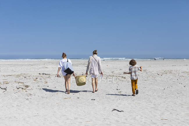 Madre e hija adolescente caminando en una playa de arena llevando una canasta, niño siguiendo. - foto de stock