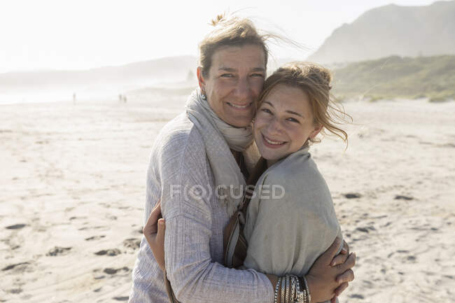 Mujer adulta y su hija adolescente abrazándose, de pie en una playa azotada por el viento - foto de stock