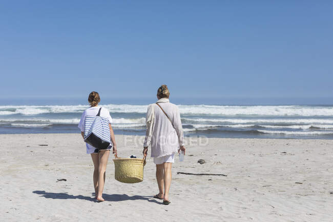 Madre e hija adolescente caminando en una playa de arena llevando una canasta - foto de stock