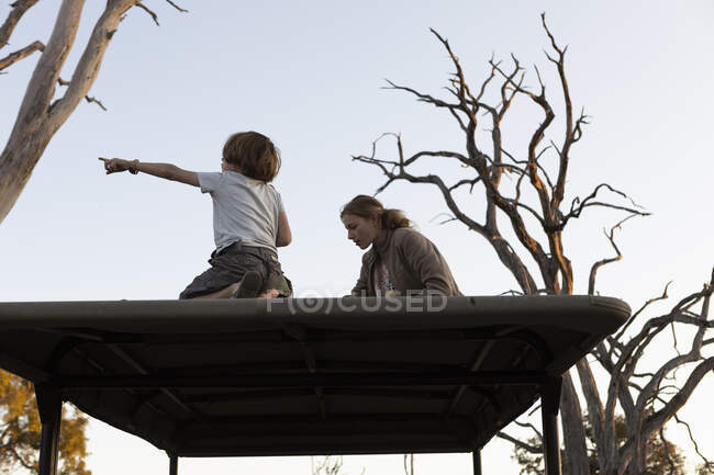 Dos niños sentados en la cima de un jeep turístico safari, en el bosque, al atardecer. - foto de stock