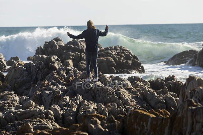 Adolescente grimpant sur les rochers déchiquetés sur une plage, de grandes vagues se brisant sur le rivage — Photo de stock
