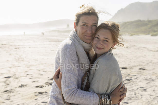 Adult woman and her teenage daughter hugging, standing on a windswept beach — Stock Photo