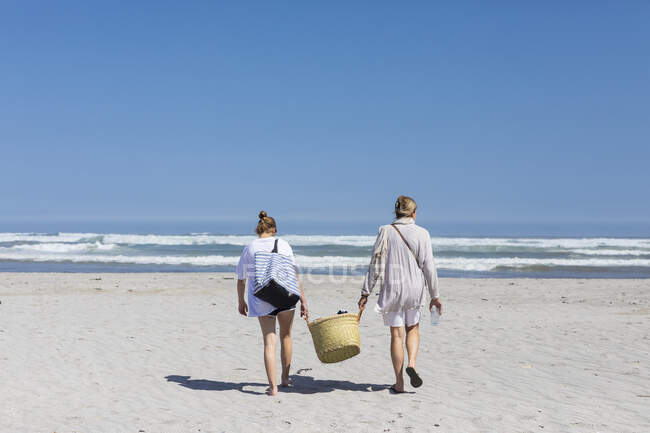 Mãe e filha adolescente andando em uma praia de areia carregando uma cesta — Fotografia de Stock